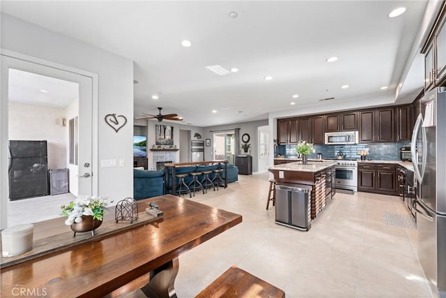 kitchen featuring backsplash, dark brown cabinets, a fireplace, stainless steel appliances, and a kitchen island with sink