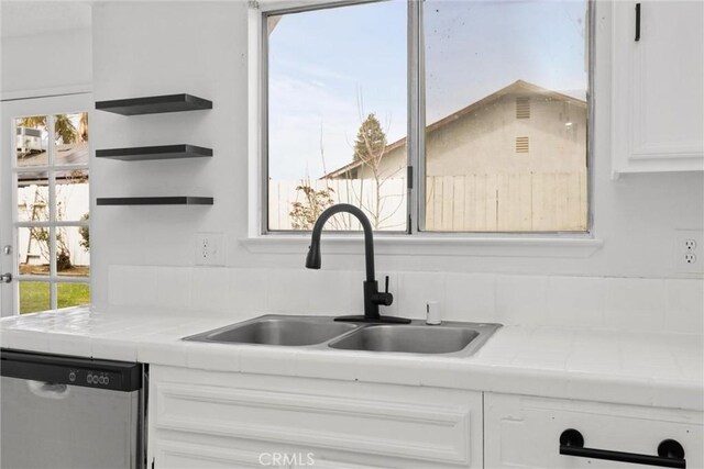 kitchen with tile counters, open shelves, stainless steel dishwasher, a sink, and white cabinetry