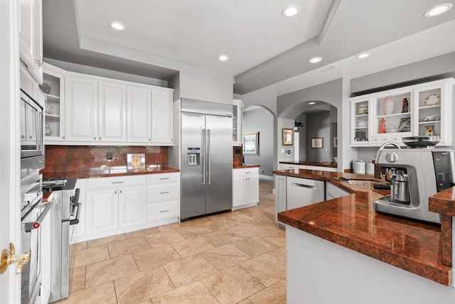 kitchen with glass insert cabinets, dark countertops, stainless steel built in refrigerator, and white cabinetry