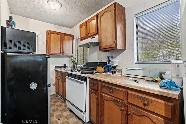 kitchen featuring brown cabinetry, light floors, freestanding refrigerator, white gas range oven, and under cabinet range hood