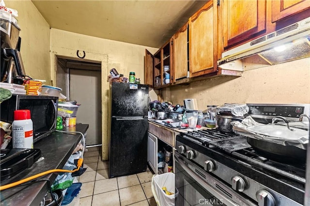 kitchen with under cabinet range hood, light tile patterned floors, brown cabinets, freestanding refrigerator, and stainless steel gas stove