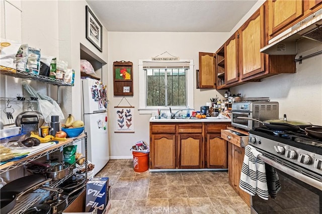 kitchen with brown cabinetry, under cabinet range hood, stainless steel gas range, and freestanding refrigerator