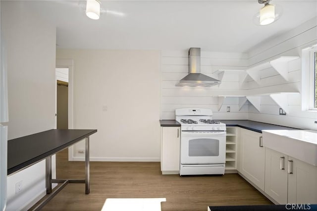 kitchen featuring wood finished floors, a sink, dark countertops, wall chimney range hood, and white gas range