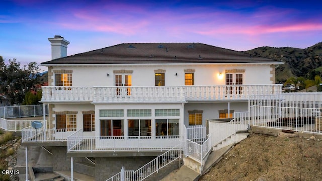 back of house at dusk featuring stairway, a balcony, a chimney, and stucco siding