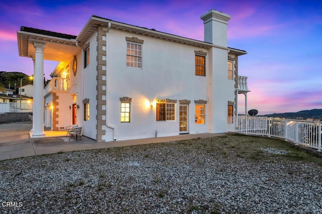 back of property at dusk with fence, stucco siding, a chimney, a balcony, and a patio area