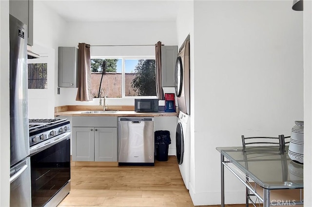 kitchen with a sink, gray cabinetry, light wood-type flooring, stacked washer and clothes dryer, and stainless steel appliances