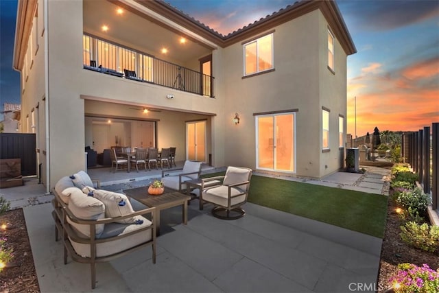 back of property at dusk featuring fence, stucco siding, outdoor lounge area, a balcony, and a patio