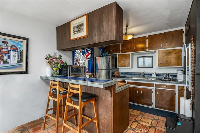 kitchen featuring a breakfast bar, a sink, freestanding refrigerator, dark brown cabinetry, and a peninsula