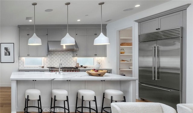 kitchen featuring stainless steel built in fridge, gray cabinets, wall chimney exhaust hood, decorative backsplash, and light stone countertops