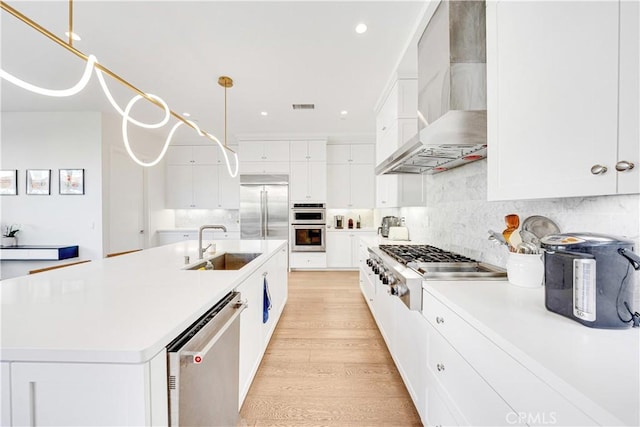 kitchen with light wood finished floors, wall chimney range hood, appliances with stainless steel finishes, white cabinets, and a sink