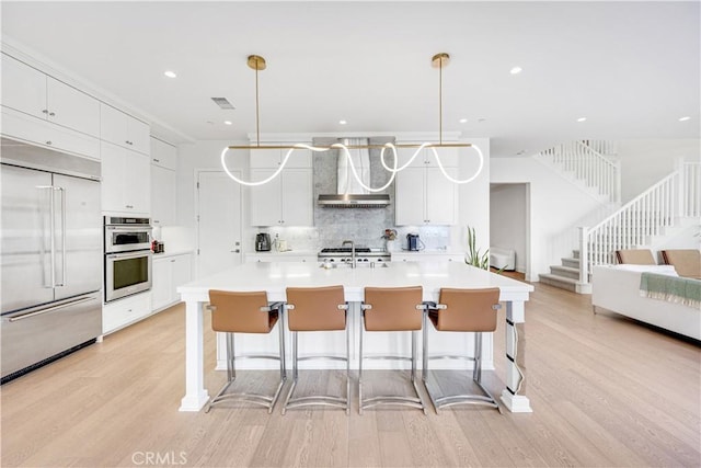 kitchen with light wood finished floors, a breakfast bar, decorative backsplash, appliances with stainless steel finishes, and wall chimney range hood