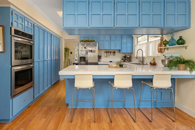 kitchen with blue cabinetry, a sink, tile countertops, stainless steel appliances, and a peninsula