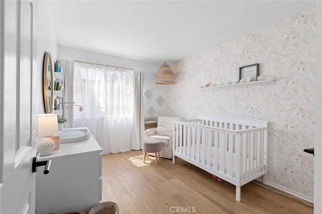 bedroom featuring light wood-type flooring, baseboards, a nursery area, and wallpapered walls