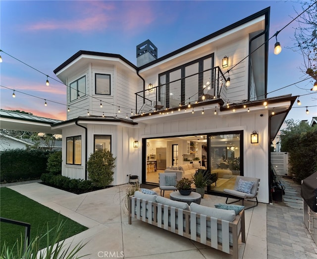 back of property at dusk featuring an outdoor living space, board and batten siding, a chimney, a balcony, and a patio