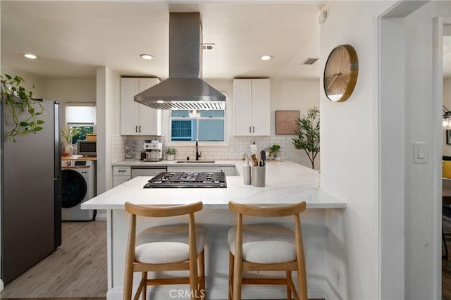 kitchen with a peninsula, island range hood, washer / clothes dryer, white cabinetry, and a sink