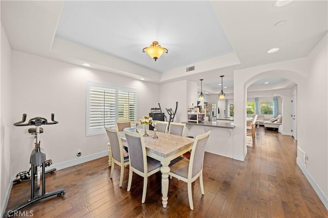 dining space featuring a raised ceiling, arched walkways, dark wood-type flooring, and baseboards