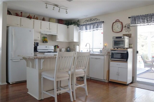 kitchen with white appliances, wood finished floors, a breakfast bar, under cabinet range hood, and white cabinetry