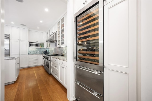 kitchen with glass insert cabinets, under cabinet range hood, wood finished floors, white cabinets, and stainless steel appliances