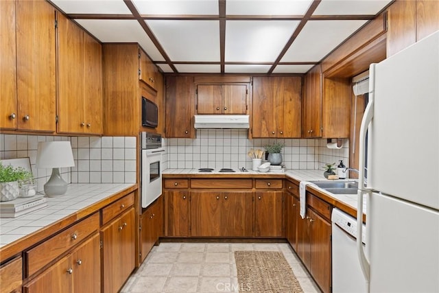 kitchen featuring brown cabinets, under cabinet range hood, a sink, white appliances, and tile counters