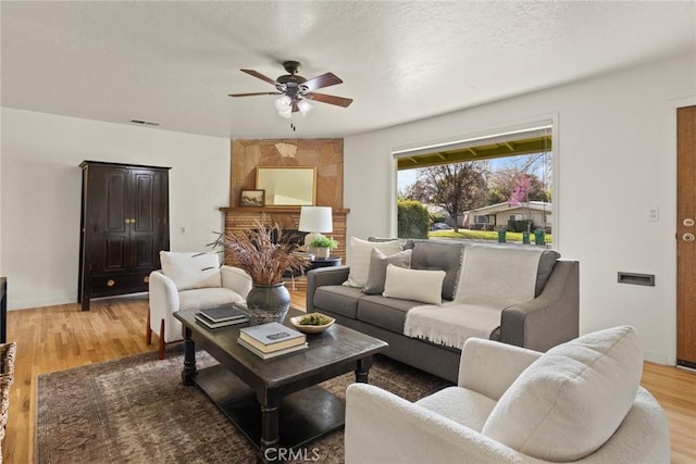 living area with light wood finished floors, visible vents, a fireplace with raised hearth, a textured ceiling, and a ceiling fan