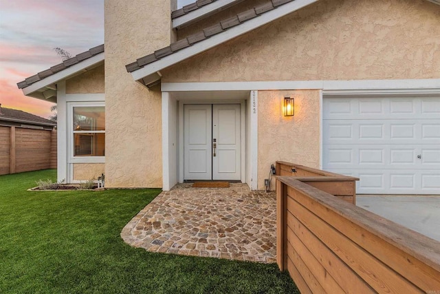 exterior entry at dusk featuring stucco siding, a lawn, a garage, and fence