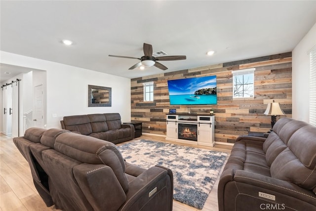 living area with a glass covered fireplace, light wood finished floors, ceiling fan, an accent wall, and a barn door