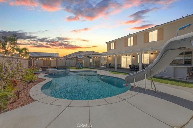 pool at dusk with a patio area, a ceiling fan, a water slide, and a fenced backyard