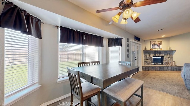 dining space with ceiling fan, baseboards, vaulted ceiling, a stone fireplace, and wood finished floors