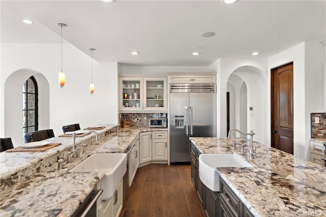 kitchen with built in refrigerator, cream cabinetry, a sink, arched walkways, and glass insert cabinets