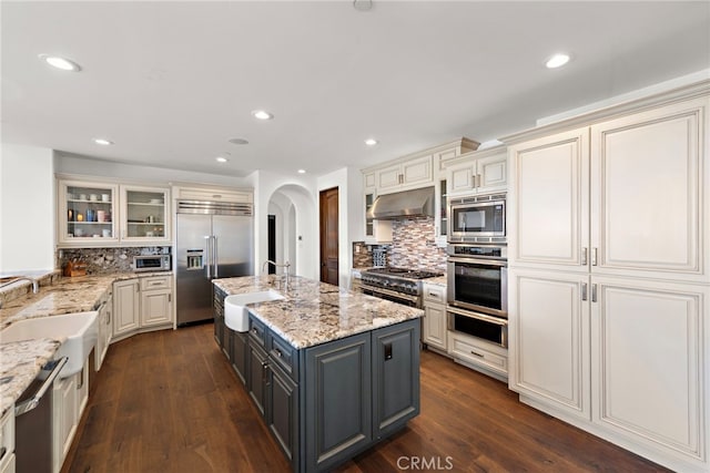 kitchen featuring gray cabinetry, under cabinet range hood, a sink, arched walkways, and built in appliances