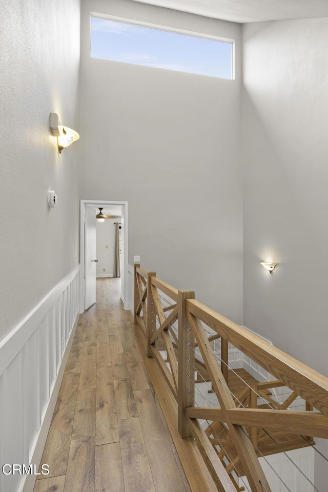 hallway featuring a wainscoted wall, light wood-style floors, a towering ceiling, a decorative wall, and an upstairs landing