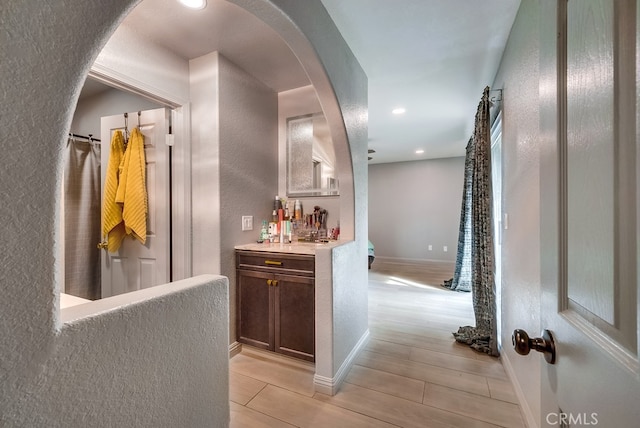 hall featuring baseboards, recessed lighting, light wood-style floors, a textured wall, and arched walkways