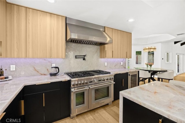 kitchen with light brown cabinets, wall chimney range hood, beverage cooler, range with two ovens, and dark cabinetry