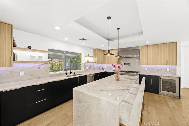 kitchen with open shelves, a raised ceiling, dark cabinetry, wall chimney exhaust hood, and a sink