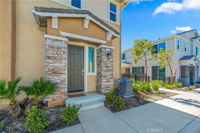 view of exterior entry featuring stucco siding, stone siding, central AC, and fence