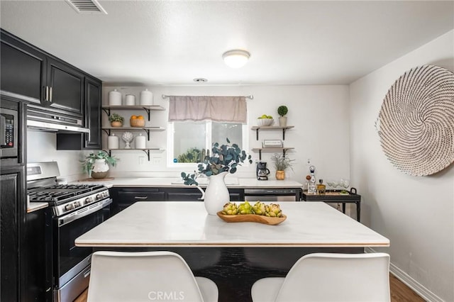 kitchen with open shelves, under cabinet range hood, stainless steel appliances, light countertops, and dark cabinets