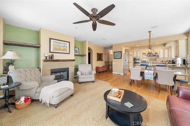 living area featuring light wood finished floors, ceiling fan with notable chandelier, a tile fireplace, and baseboards