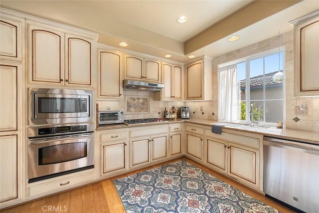 kitchen featuring under cabinet range hood, appliances with stainless steel finishes, light wood-style floors, cream cabinetry, and a sink