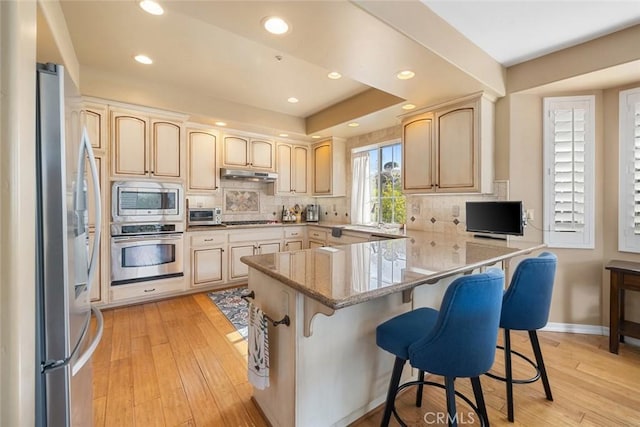 kitchen featuring light stone counters, light brown cabinetry, under cabinet range hood, appliances with stainless steel finishes, and light wood-type flooring