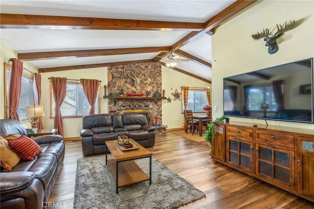 living area with a ceiling fan, a stone fireplace, wood finished floors, and vaulted ceiling with beams