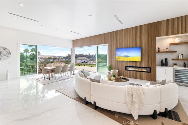 living room featuring beverage cooler, wood walls, recessed lighting, a glass covered fireplace, and marble finish floor