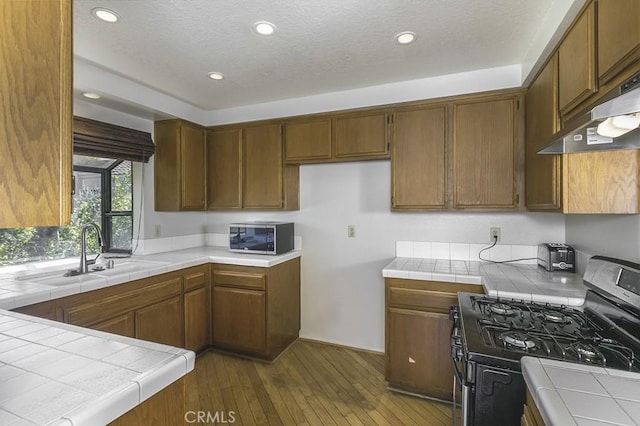 kitchen featuring tile counters, dark wood finished floors, under cabinet range hood, stainless steel appliances, and a sink