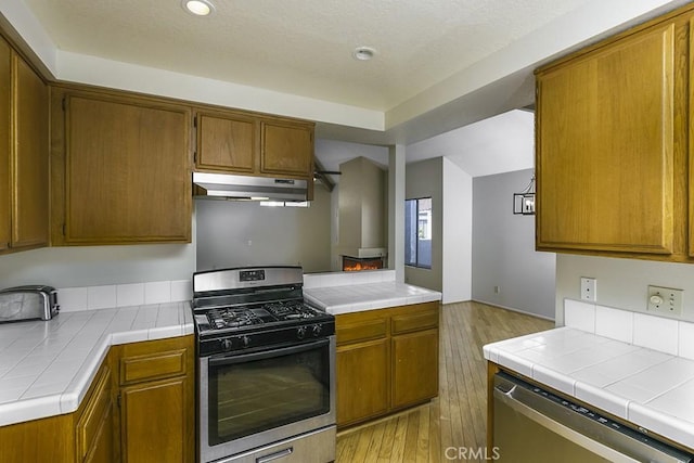 kitchen featuring brown cabinetry, light wood finished floors, tile counters, under cabinet range hood, and appliances with stainless steel finishes