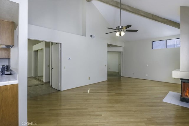 unfurnished living room featuring visible vents, a wood stove, light wood-style flooring, a ceiling fan, and vaulted ceiling with beams