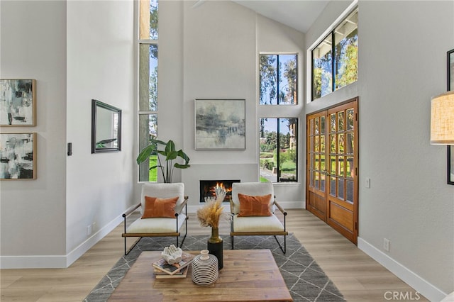 sitting room with high vaulted ceiling, a healthy amount of sunlight, wood finished floors, and a warm lit fireplace