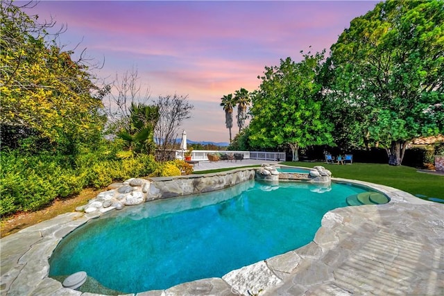 pool at dusk with a patio, a lawn, fence, and a pool with connected hot tub