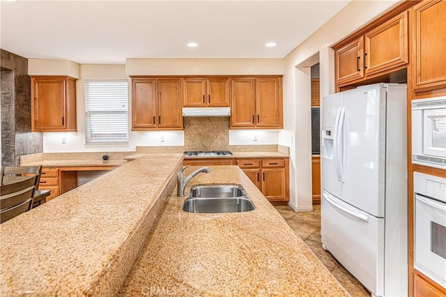 kitchen featuring white appliances, brown cabinetry, under cabinet range hood, and a sink