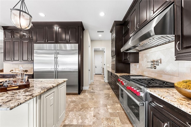 kitchen featuring stone finish floor, under cabinet range hood, premium appliances, light stone counters, and dark brown cabinetry