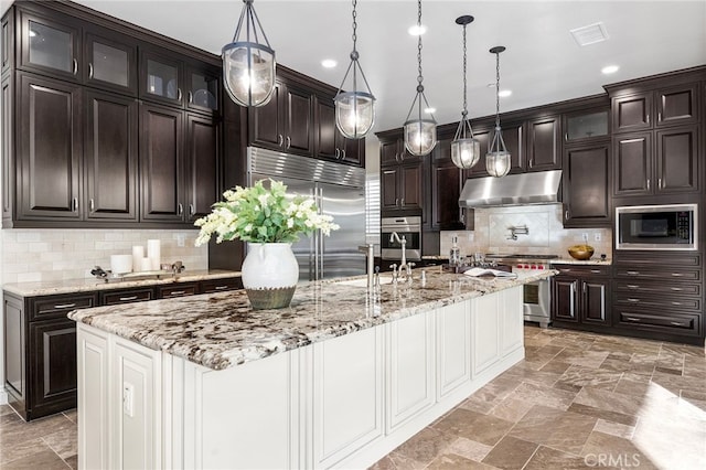 kitchen with visible vents, an island with sink, under cabinet range hood, stone finish flooring, and built in appliances