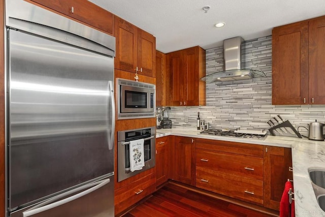 kitchen with built in appliances, wall chimney exhaust hood, dark wood-type flooring, and decorative backsplash